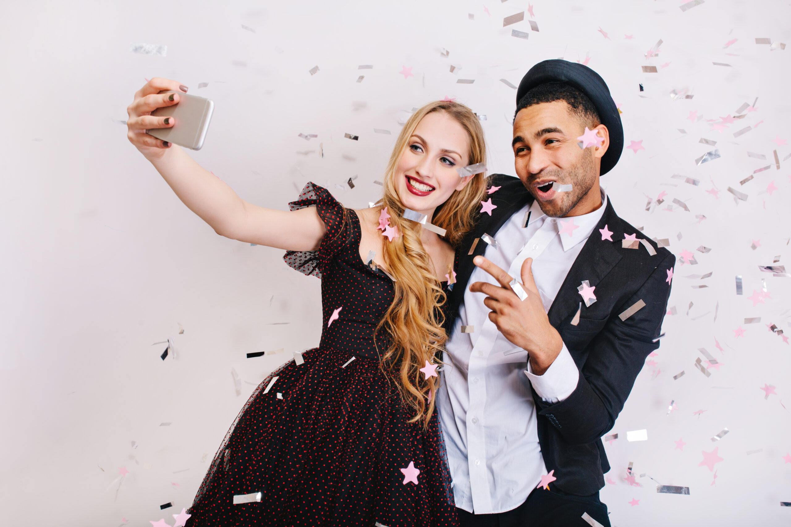 Attractive young woman with long blonde hair in luxury evening dress making selfie in tinsels with joyful handsome man on white background. Celebrating party, Valentine s day, lovers